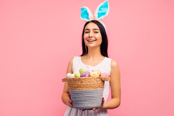 Happy woman in rabbit ears holding a basket with colored eggs, a traditional holiday, pink background, Easter mood