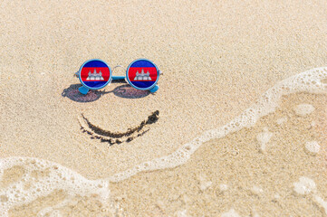 Fototapeta na wymiar A painted smile on the sand and sunglasses with the flag of the Cambodia. The concept of a positive and successful holiday in the resort of Cambodia.
