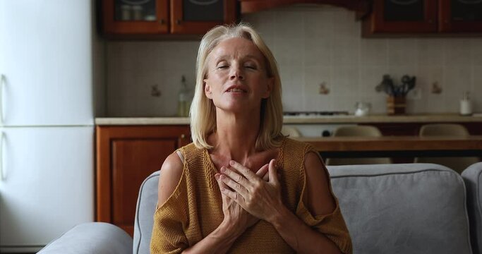 Middle-aged woman closing eyes sincere begging, praying seated on sofa at home, put folded arms on chest asks higher forces for help, luck or miracle. Faith, religion, express gratitude to God concept
