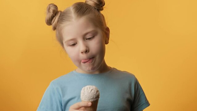 Happy toothless girl eating vanilla ice cream on yellow background. Child with ice cream looking at camera. Close-up in 4K, UHD
