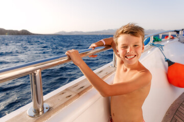 Young boy standing on a deck of yacht closing eyes and enjoying the trip in sunny day. Kid is wearing shorts 