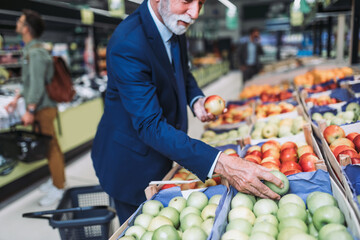 Senior bearded man buying fruits at the supermarket. Other customers in background. Consumerism concept.