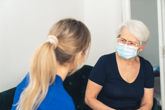 Blonde Nurse In A Blue Uniform Has A Home Visit. Elderly Masked Lady Complaining About Health Problems. High Quality Photo