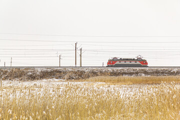 A lone electric train without wagons moves along the railway past yellow grass against the backdrop of white snow.