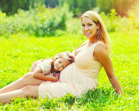 Pregnancy and family concept, happy pregnant woman, little child daughter touches belly mother on the grass in summer day