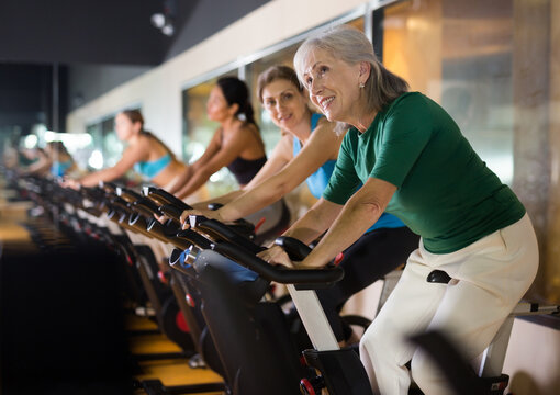 Positive mature female riding exercise bike during cycling class in modern gym