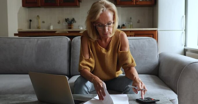 Older woman sits on couch use calculator counts expenses and incomes, makes payment use e-bank app on laptop, pay monthly household bills. Budget control, net worth, manage personal financial concept