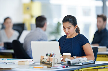 Immersed in her work. Shot of a designer sitting at her desk working on a laptop with colleagues in the background.
