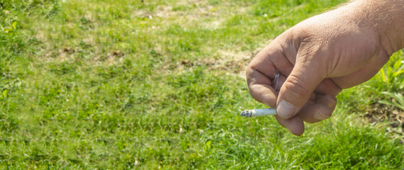 Close-up of a man's hand smoking a cigarette, on a grass background, outdoors, smoking cessation concept, space for text on the left