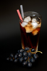 Refreshing grape juice with ice in a tall glass and a bunch of ripe black grapes on a dark background.