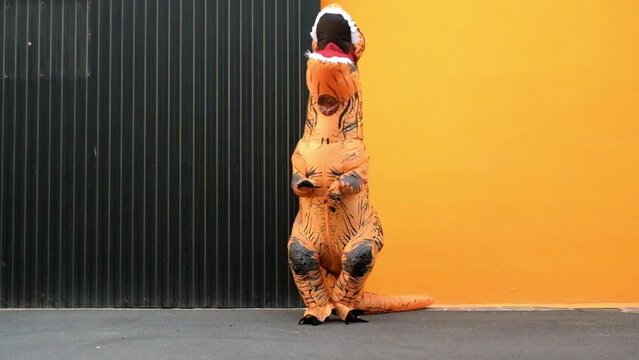 One happy and funny dinosaur costume dancing in the street with a orange colorful background - t-rex having fun - funny man inside of a costume of dino
