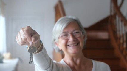 Close up of old and mature woman at home holding keys of house to the camera. Portrait of female senior smiling and looking at the camera.
