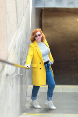 Fototapeta premium A young active woman with red hair, in bright clothes, a yellow coat and jeans
