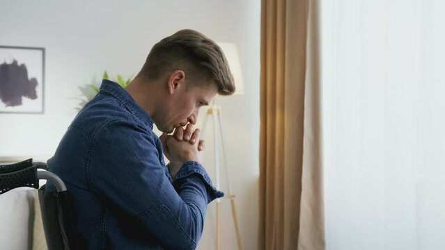Zoom in portrait of hopeless middle aged man with disability praying to God, sitting in wheelchair against window