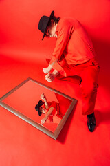 Tall handsome man dressed in red shirt and black hat sitting on the red background and looking into the mirror.