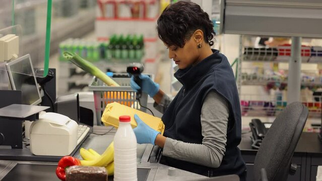 Young female cashier selling products and smiling while sitting at checkout of store spbi. 4k Close view of american african woman takes goods in hand and scans with device, looks with smile and sits