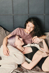 Attractive young lesbians lying on bed and touching hands of each other while looking into eyes