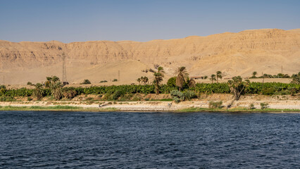 Palm trees and green vegetation grow on the banks of the Nile. Power towers are visible. A high sand dune against a clear sky. Ripples on the blue water. Egypt