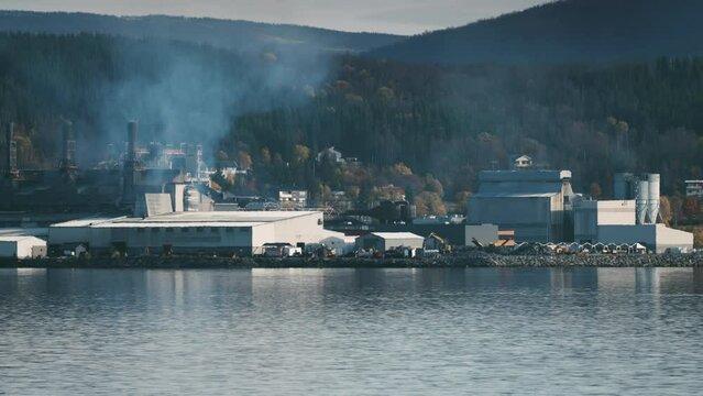 A working ferrosilicon production on the shore of the fjord. Smoke and steam are rising from the chimneys. Crane unloading goods. Heavy machines and excavators move on the site.