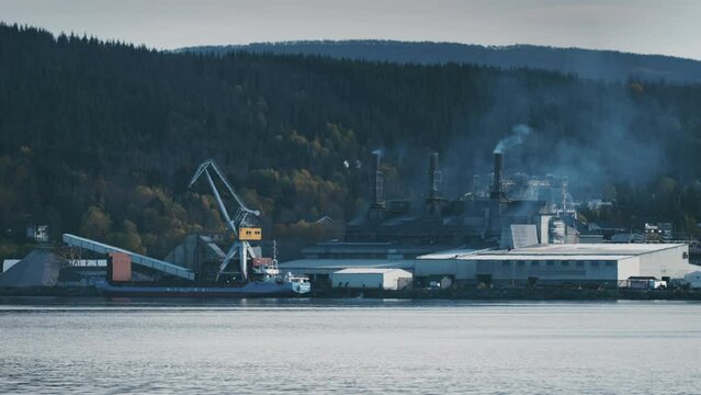 A ferrosilicon production plant on the shore of the fjord. Smoke and steam are rising from the chimneys. Crane unloading goods.