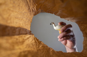 A hand places a goose figurine in a paper bag. A mature man's hand holds a child's toy over an open brown bag.  Shot from the bottom up. Close up. Selective focus.