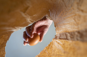 A hand places a chicken egg in a paper bag. A mature man's hand holds a brown egg over an open brown bag. Shot from the bottom up. Close up. Selective focus.