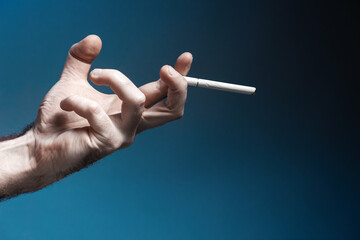 World No Tabacco day. A pale, crooked male hand, close-up, holding a smoking cigarette. Dark blue background. Copy space. The concept of nicotine addiction