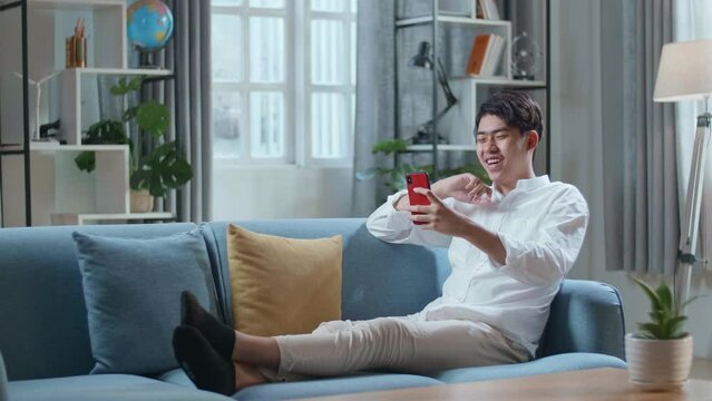 Full Body Of Smiling Asian Man Having Video Call On Smartphone While Lying On Sofa In The Living Room 
