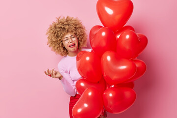 Upset crying woman has spoiled makeup curly hair feels dejected holds bunch of heart balloons keeps hand raised expresses negative emotions isolated over pink background. Festive event concept
