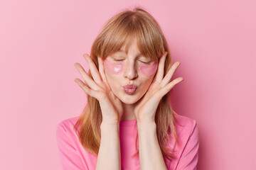 Red haired freckled girl keeps lips folded has freckled skin applies hydrogel patches under eyes...