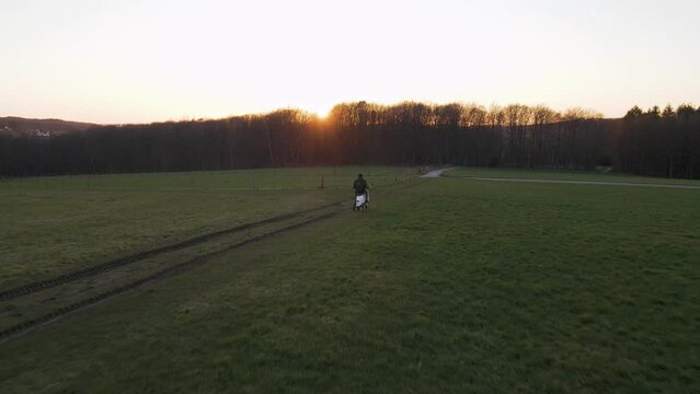 Young man sitting on a scooter within a lush, green field during a magnificent sunset in Germany. Aerial wide angle fly-over shot