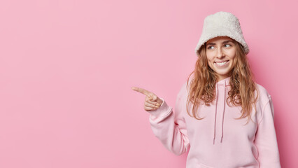 Beautiful young woman with wavy hair smiles happily points left with index finger advertises...