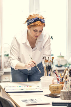 A young active female artist, with red hair, in a white shirt and jeans with a bandage on her head, paints standing at the table