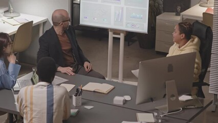 Medium slowmo of team of investment analysis specialists discussing financial data on monitor while sitting together at desk in modern office at night - Powered by Adobe
