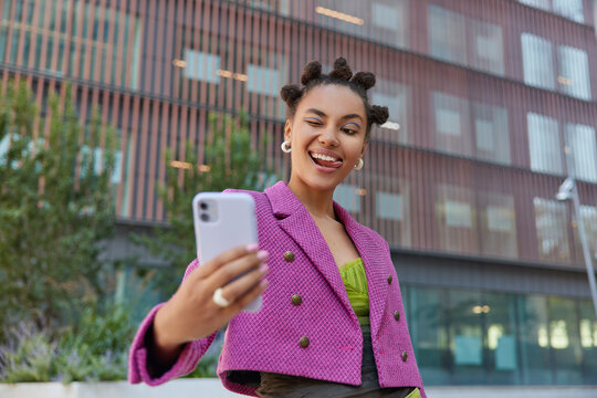Fashionable young woman with bun hairstyle winks eye poses for selfie at smartphone camera wears fashionable pink jacket poses outdoors in urban setting has cheerful expression. Street lifestyle