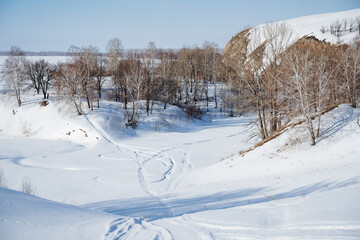 Fototapeta na wymiar Snowmobile tracks in the snow, a winter forest, the river valley below is covered with snow, a descent from the mountain, a path through the forest along a ravine.
