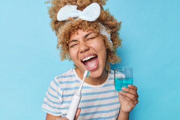 Tooth care concept. Positive young woman with curly combed hair holds electric toothbrush and glass...