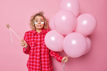 Fototapeta na wymiar Scared terrified woman holds magic wand and bunch of inflated balloons cannot believe her eyes feels fearful applies beauty patches under eyes stands against pink background. Birthday party.