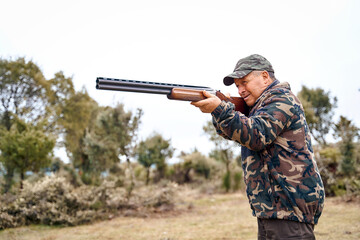 Senior male hunter in camouflage clothing in the field aiming hunting shotgun during a hunt. High quality photo
