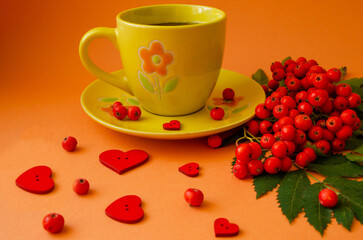 Yellow mug with a saucer and ripe rowan berries and red hearts on an orange background. Cozy autumn concept