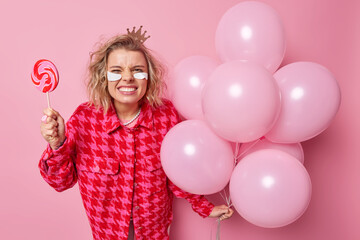 Fototapeta premium Blonde pretty European woman smirks face clenches teeth holds caramel candy and bunch of inflated balloons wears crown checkered jacket beauty patches under eyes isolated over pink background