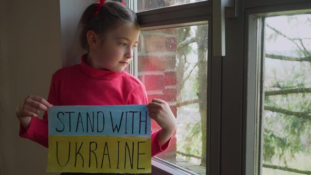 Girl looking outside window and holding banner with inscription Stand with Ukraine at blue yellow flag. Crisis, peace, stop aggression in whole world