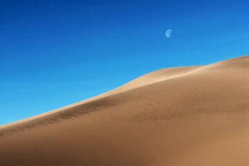 Fototapeta na wymiar Moon against blue sky over sand dunes. Sand dunes against the blue sky.