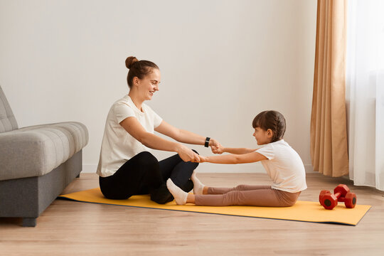 Image of woman with little girl wearing sportswear doing sport exercises at home, sitting on floor, holding hands together and practicing yoga at home, doing stretching exercise.