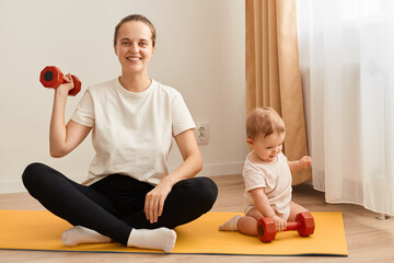 Fototapeta na wymiar Portrait of adorable athletic woman wearing white t shirt and black leggins doing sport exercises with baby nearby, raising her arm with dumbbell, having workout for biceps.