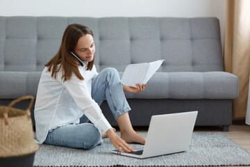 Image of dark haired young adult woman wearing white shirt and jeans sitting on floor near sofa,...