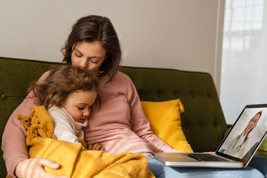Mother embracing sick daughter consulting doctor online through laptop at home