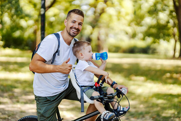 A father giving thumbs up for a healthy living while sitting with son on a bike in nature.