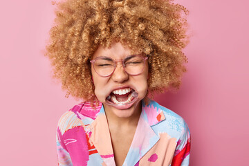 Headshot of curly haired woman with expander in mouth shows white teeth keeps eyes closed wears spectacles and colorful shirt poses against pink background. Emotional female uses dental retractor