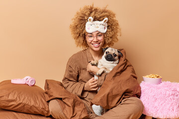 Obraz premium Happy female pet owner with cute pug dog wrapped in blanket wears pajama and sleepmask plays with favorite pet pose in bedroom against brown background. Domestic atmosphere animals friendship